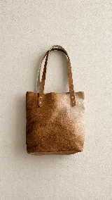 Bolso tipo tote confeccionado artesanalmente en cuero vacuno con pelo, presentando un diseño degradado en tonos marrones y beige. Cuenta con dos asas largas de cuero liso sujetas con remaches metálicos.