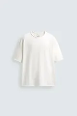 Camiseta de corte relajado (relaxed fit) color blanco roto, con cuello redondo y manga corta. Presenta un bordado sutil en el pecho con diseño floral en tonos claros.