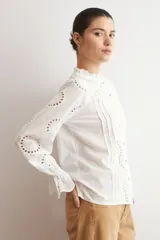 Camisa blanca de manga larga con cuello alto y volados, confeccionada en tela con bordado inglés y detalles calados verticales. Presenta puños con volados y cierre frontal con botones.