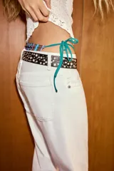 Cinturón de efecto piel en color negro, con diseño de tachas metálicas y perforaciones decorativas a lo largo de toda la pieza. Cuenta con hebilla metálica cuadrada en tono plateado.