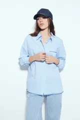 Camisa de silueta amplia y caída fluida, color celeste con rayas verticales blancas. Presenta cuello camisero clásico, cierre frontal con botones y un bolsillo aplicado en el pecho. Las costuras están resaltadas en azul oscuro.