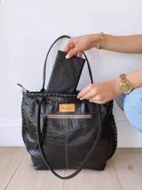 Cartera tipo tote de cuero negro con textura de cocodrilo. Presenta detalles de costuras trenzadas a lo largo de la estructura y tiras decorativas laterales. Cuenta con doble asa de hombro y cierre superior.