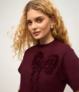 Remera femenina de manga corta color bordó, con cuello redondo y una aplicación figurativa en forma de lazo en la parte delantera, hecha en tejido fruncido que crea relieve. Confeccionada en algodón certificado BCI.