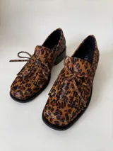 Mocasín de cuero marrón con textura granulada, diseño de horma cuadrada, detalle de flecos y lazo decorativo en el empeine, montado sobre una suela y taco bajo de color negro.