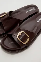 Sandalia plana de eco cuero negro, con tira ancha en el empeine decorada con una hebilla metálica dorada cuadrada y suela dentada.