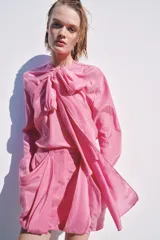 Blusa fluida de color rosado con cuello redondo ajustable mediante lazadas del mismo tejido, manga larga con puño y bajo asimétrico con aberturas laterales.