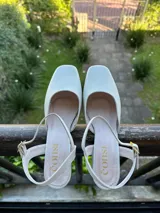 Zapatos cerrados de cuero natural color blanco marfil, con diseño inspirado en bailarinas de ballet, punta cuadrada, taco de 4 cm y tira ajustable en el talón con hebilla metálica dorada.