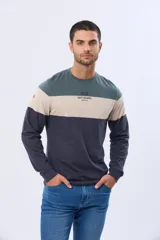 Remera de manga larga con cuello redondo y diseño de rayas horizontales en azul verdoso, beige y azul marino. Presenta una estampa frontal con texto y un parche decorativo en la manga.