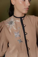 Chaqueta gris de silueta femenina con mangas abullonadas, cuello redondo y cierre frontal con botones forrados. Presenta detalles de bordado artesanal a mano en los hombros y solapas.