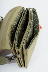 Bandolera de diseño rectangular en color verde oliva, con solapa frontal y logo de la marca grabado. Incluye una correa larga ajustable con el nombre de la marca tejido y un charm decorativo con forma de corazón y letra R en color rojo.