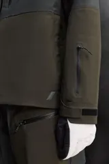 Cazadora deportiva de esquí con diseño bicolor en tonos negro y verde oliva. Presenta capucha amplia con visera, cierre frontal con cremallera termosellada, bolsillos frontales y en manga con cierres impermeables, y aberturas de ventilación en las axilas. Incluye faldón interior antinieve, puños ajustables y tecnología de rescate RECCO.