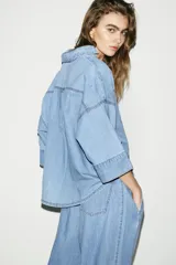 Camisa de denim en tono azul claro, con cuello solapa, escote en pico y cierre frontal con botones. Presenta mangas anchas por debajo del codo y un diseño de bajo asimétrico.