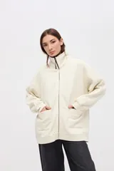 Campera oversize de felpa con friza, color crudo, con cierre metálico frontal, cuello alto y bolsillos laterales amplios. Presenta detalles de cortes en mangas y hombros, y bordado decorativo.