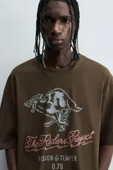 Camiseta de algodón color marrón oscuro, corte relajado (relaxed fit) y manga corta. Presenta un estampado frontal con un lobo o cánido corriendo, el texto "The Riders Project", "VISION & TEMPER" y "0.79 per gallon" en tonos grises y rosados.