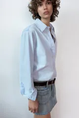 Falda mini de denim en tono azul claro, con tiro medio, trabillas para cinturón y bolsillos de plastrón en el frente. Presenta cierre frontal con cremallera y botones metálicos.