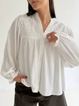 Blusa blanca de corte holgado con cuello mao, botones frontales y detalles bordados en el pecho. Presenta mangas largas con puños con volados.