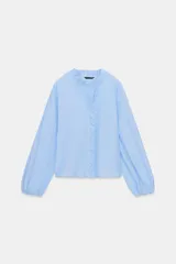 Blusa celeste de popelín, con cuello subido y escote de pico, manga larga abullonada terminada en elástico, y detalles de volantes en el cuello y la botonadura frontal con cierre de botones ocultos.