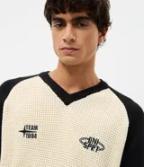 Suéter masculino de punto en modelo boxy, con cuello en V, manga larga y diseño bicolor en beige y negro. Presenta inscripciones bordadas en el pecho, incluyendo la palabra 'Untitled' en tipografía cursiva, y detalles de rayas en los puños, las mangas y el ruedo.