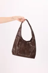 Bolso tipo hobo de símil cuero color marrón, con detalle de tachas metálicas plateadas en el bolsillo frontal y a lo largo de la manija.