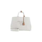 Cartera Prada tipo shopper de lona color beige claro, con asas superiores, detalles de cuero marrón y correa de hombro ajustable con el logo de la marca bordado. Cuenta con el nombre y logo de Prada bordados en el frente y un llavero colgante de cuero.
