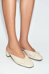 Zapatos de tacón tipo bailarina destalonada en piel color beige, con detalle de lazo en la parte delantera, tacón en bloque y punta redonda.