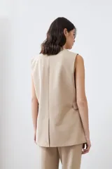 Chaleco largo de corte sastrero en color beige, con escote en V profundo, cierre frontal con un botón metálico y bolsillos laterales con solapa.