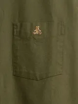 Camisa de corte clásico confeccionada en tejido Oxford, color verde oliva. Presenta cuello abotonado, cierre frontal con botones, mangas largas con puños abotonados y un bolsillo en el pecho con el logo del oso Brannan bordado.