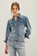 Camisa de jean de corte clásico con efecto lavado, cuello camisero, cierre frontal con botones metálicos y bolsillos frontales con solapa. Presenta mangas largas con diseño abullonado en los hombros y puños abotonados.