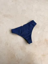 Bombacha de bikini tipo vedetina, confeccionada en tela brillante color azul. Presenta una argolla decorativa de madera en uno de los laterales y cuenta con forro interno.