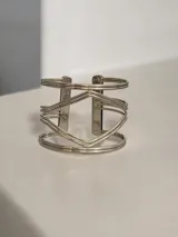 Brazalete abierto de diseño geométrico en tono plateado, compuesto por múltiples líneas metálicas que forman una estructura central en forma de rombo.