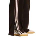 Pantalon deportivo Adidas de corte holgado, color marrón con las icónicas tres tiras laterales en tono claro y logo del trébol bordado en la pierna. Cuenta con cintura elástica y bolsillos laterales con cierre.
