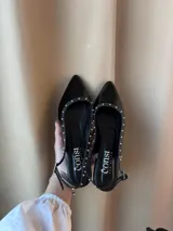Chatitas destalonadas de cuero charolado negro, con punta fina y tachas metálicas plateadas decorativas en los bordes. Cuentan con una tira ajustable en el talón y un tacón bajo.