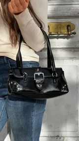 Cartera de hombro negra con acabado brillante, diseño rectangular con solapa frontal, cierre de hebilla metálica y detalles de tachas plateadas en los extremos inferiores.