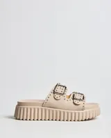 Sandalias tipo mule de color beige, con suela de plataforma texturizada. Posee dos tiras anchas en el empeine con hebillas metálicas cuadradas y decoración de remaches plateados.