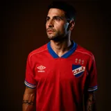 Camiseta de fútbol Umbro del Club Nacional de Football, versión jugador. Diseño en color rojo con cuello en V azul, detalles tricolores en los puños de las mangas y escudo del club con tres copas bordadas en el pecho.
