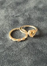 Set de dos anillos de plata 925 con acabado brillante. El primer anillo presenta un diseño entrelazado con pequeñas incrustaciones de zirconias, mientras que el segundo anillo destaca por un dije central en forma de corazón con una piedra amarilla y borde de zirconias.