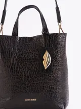 Tote bag Jazmin Chebar de cuero vacuno negro con textura grabada. Presenta un diseño compacto con doble asa superior y un tag colgante con un aplique metálico en forma de labios.