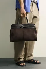 Bolso de mano estructurado en color marrón, confeccionado en una combinación de materiales que incluye gamuza en el cuerpo principal y cuero en la parte superior y asas. Presenta un detalle de cinturón decorativo con hebilla metálica plateada en la parte frontal, doble asa de mano y cierre principal con imán.