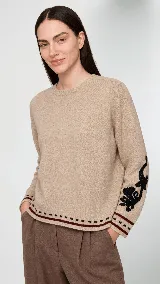 Sweater de lana color beige con calce holgado y cuello redondo. Presenta un bordado negro con diseño de arabescos en una de las mangas y detalles de rayas y puntos en contraste en el ruedo y los puños.
