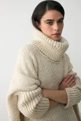 Poncho de tejido artesanal en color crudo, con cuello alto tipo polera, mangas largas y terminaciones con flecos en el ruedo.