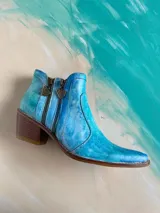 Bota corta de cuero vacuno pintada a mano en tonalidades turquesa y celeste, con taco cuadrado de madera, doble cierre lateral con tiradores metálicos personalizados y punta fina.