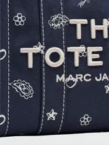 Cartera tipo tote bag de lona azul marino con estampado de motivos blancos y letras blancas bordadas en el frente. Cuenta con doble asa superior y cierre superior con cremallera.
