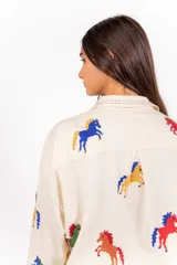 Camisa de corte clásico y calce amplio, color marfil, con cuello camisero, mangas largas y cierre frontal con botones. Presenta bordados coloridos de caballos en contraste distribuidos en la prenda.