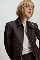 Chaqueta corta de denim oscuro con cuello solapa, cierre frontal con cremallera y bolsillos delanteros de plastrón con solapa. Presenta mangas largas con hombreras y costuras a la vista.