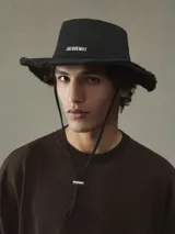 Sombrero de expedición Jacquemus modelo Le Bob Artichaut de color negro, confeccionado en algodón. Presenta ala ancha deshilachada, pespuntes decorativos, cordón ajustable con tope metálico y el logo de la marca en letras metálicas plateadas en el frente.