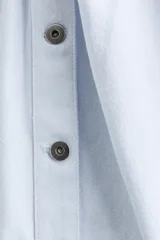 Camisa corta de sarga de algodón en color celeste, con cuello elevado, botonadura frontal con botones oscuros y corte trapecio. Presenta hombreras, mangas largas abullonadas con puños abotonados y detalles de frunces en los hombros.