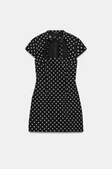Vestido corto negro con estampado de lunares blancos, cuello redondo con lazada y escote cuadrado. Cuenta con mangas cortas y cierre trasero con cremallera oculta.