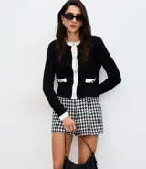 Cardigan de punto negro con detalles en blanco en el cuello, la botonera y los bolsillos frontales. Presenta un diseño corto, manga larga y cierre frontal con botones perlados.