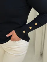 Polera negra de cuello alto y manga larga, con detalle de botones dorados en los puños. Diseño ajustado al cuerpo con tejido de punto fino.