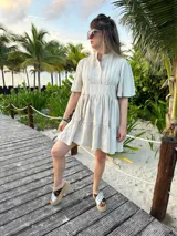 Vestido corto color beige de lino, con cuello mao, mangas cortas con volados, tablas en el canesú y falda escalonada con volantes.